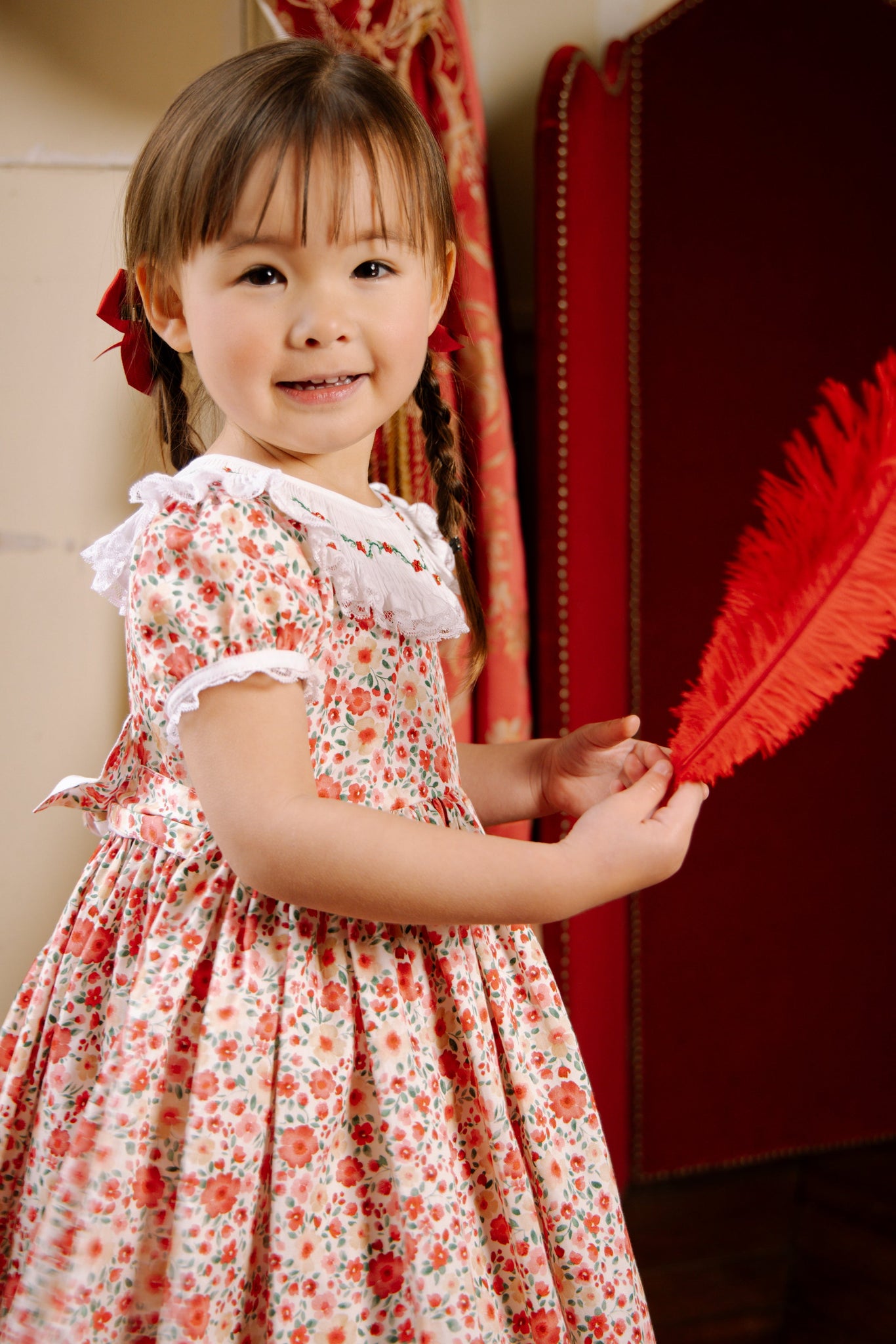 Young girl in Celestine Red Floral Smocked Dress holding a red feathered object indoors.