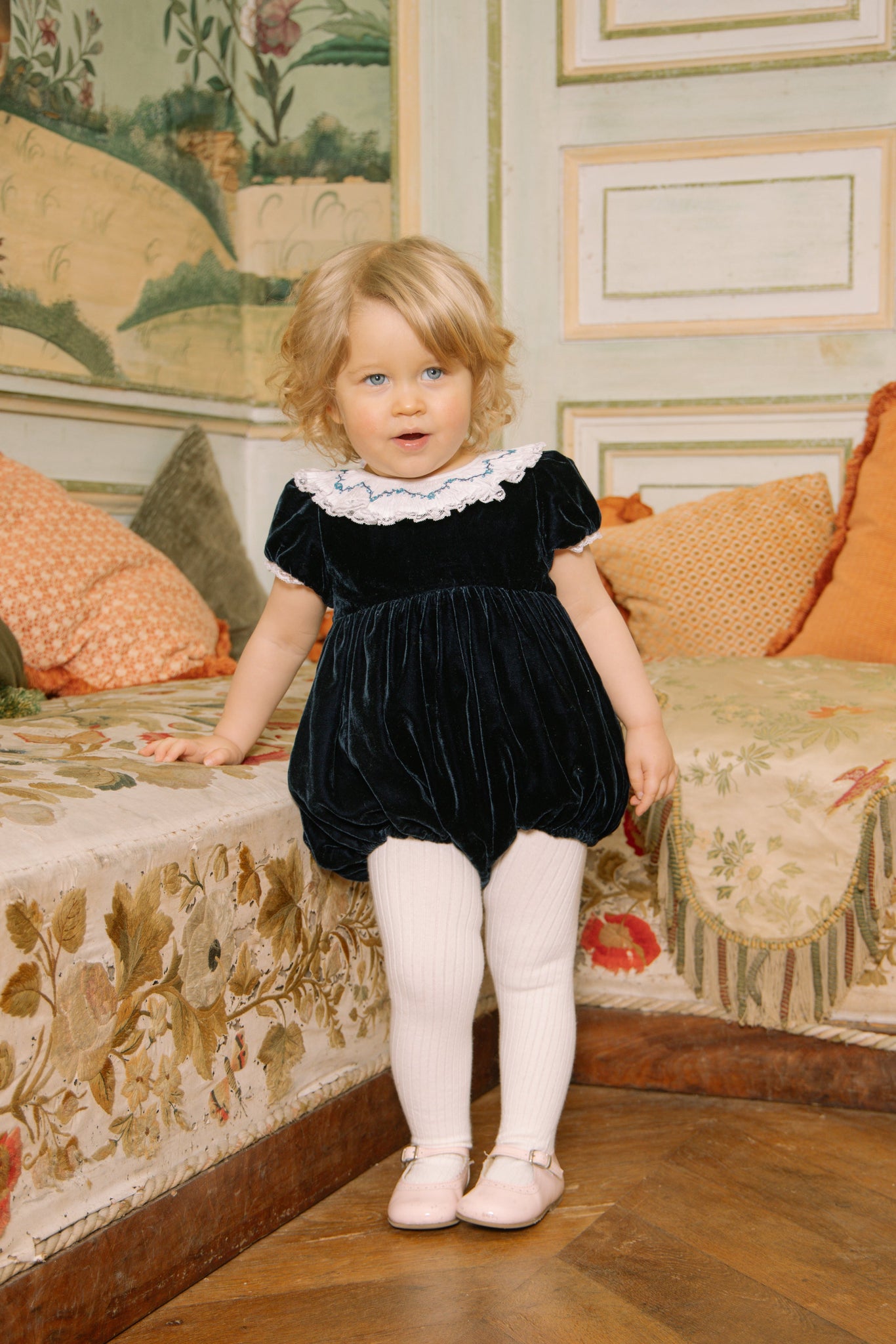 Young child in Celestine Navy Velvet Smocked Romper standing in a decorated room with floral patterns.