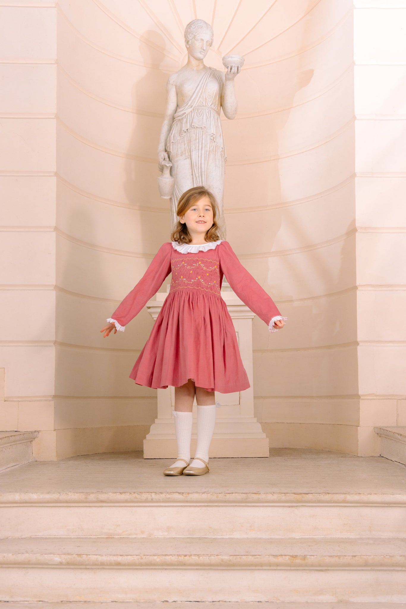 Young girl in Chiara Pink Smocked Dress standing on steps with a classical statue in the background