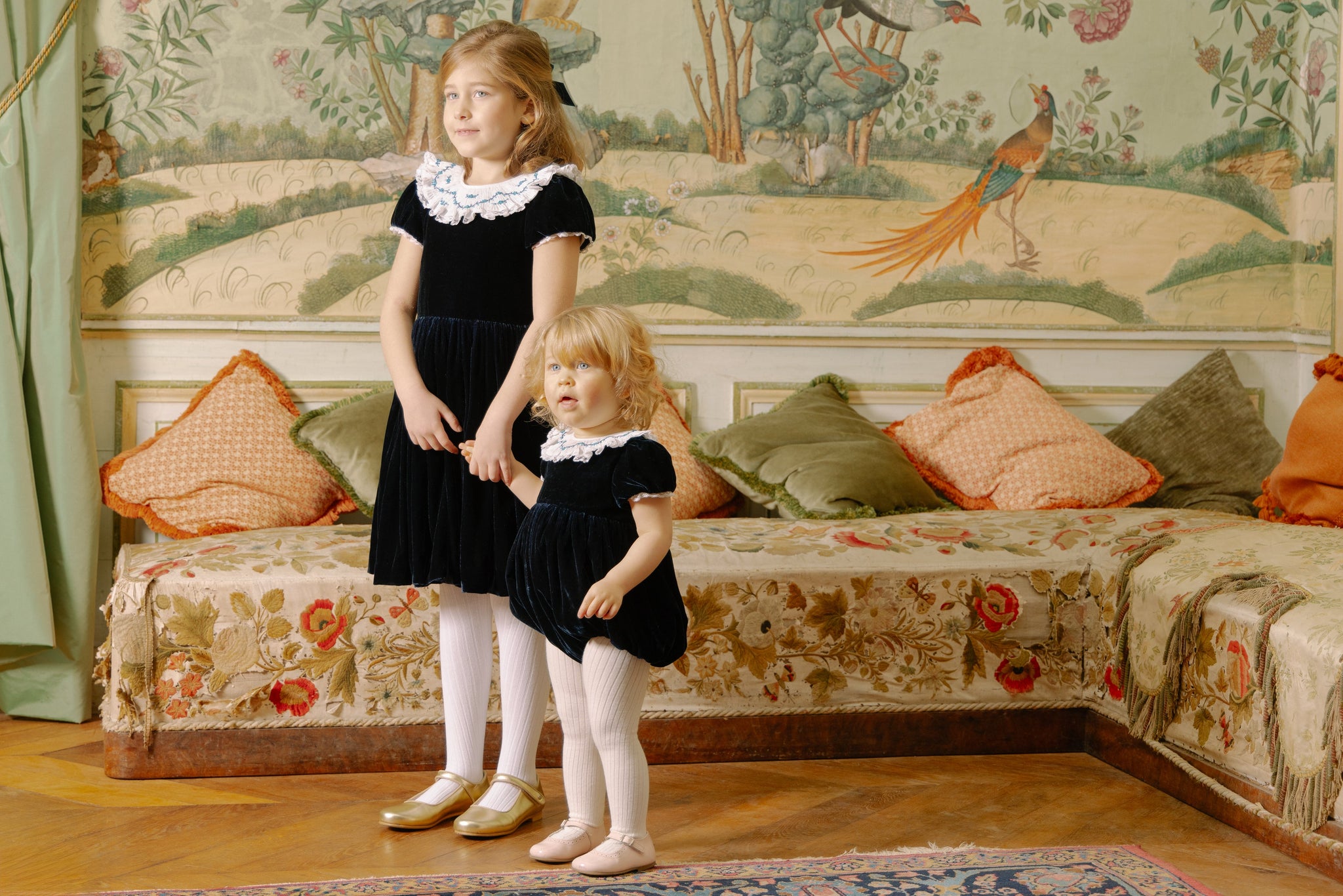 Two young girls standing in a room with decorative wallpaper and furniture.