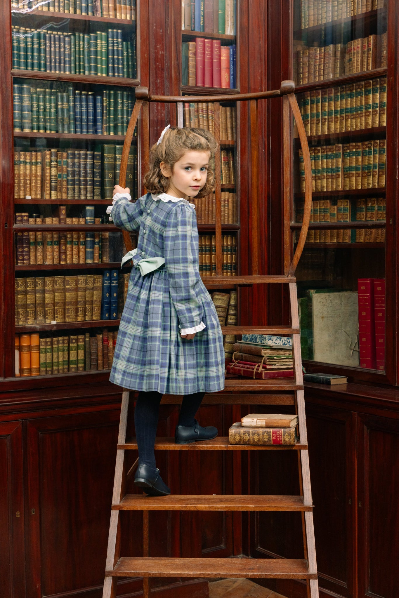 Young girl standing on a wooden ladder in a library filled with books.