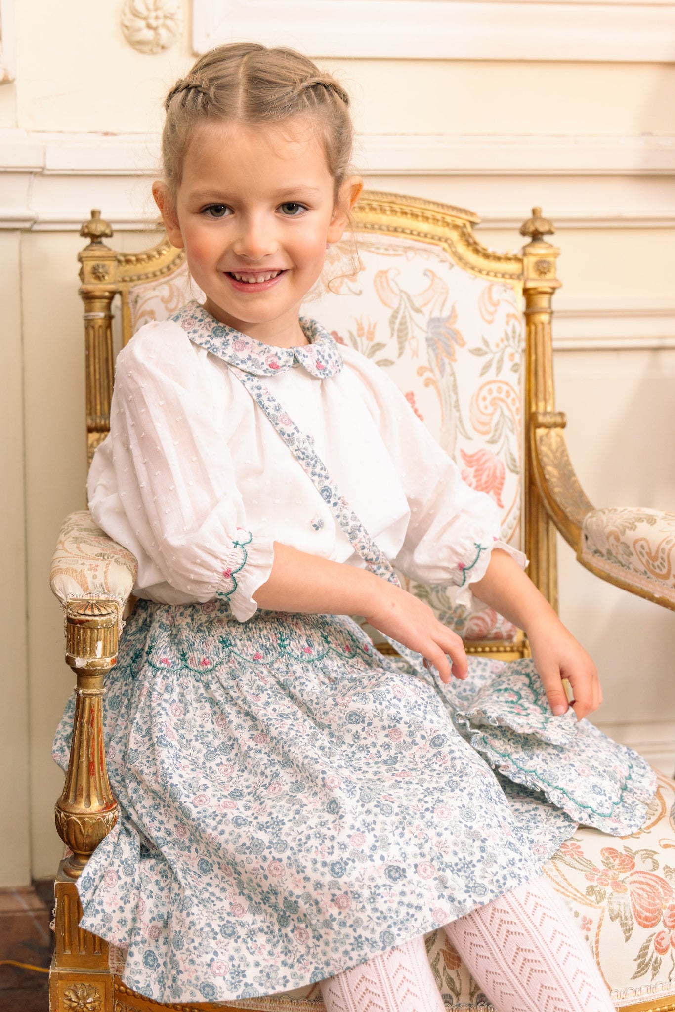 A young girl sitting on a chair, wearing Marie Blue Floral Smocked Skirt Blouse Set