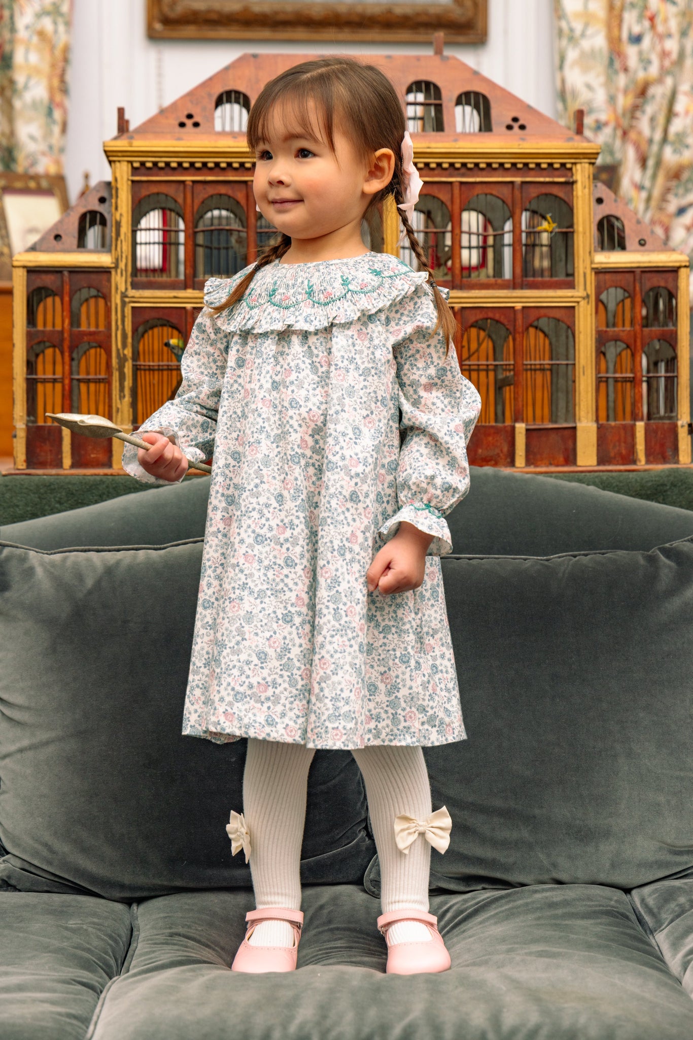 Child in Yse Blue Floral Long Sleeve Bishop Dress and Purse standing on a green couch with a wooden toy house in the background