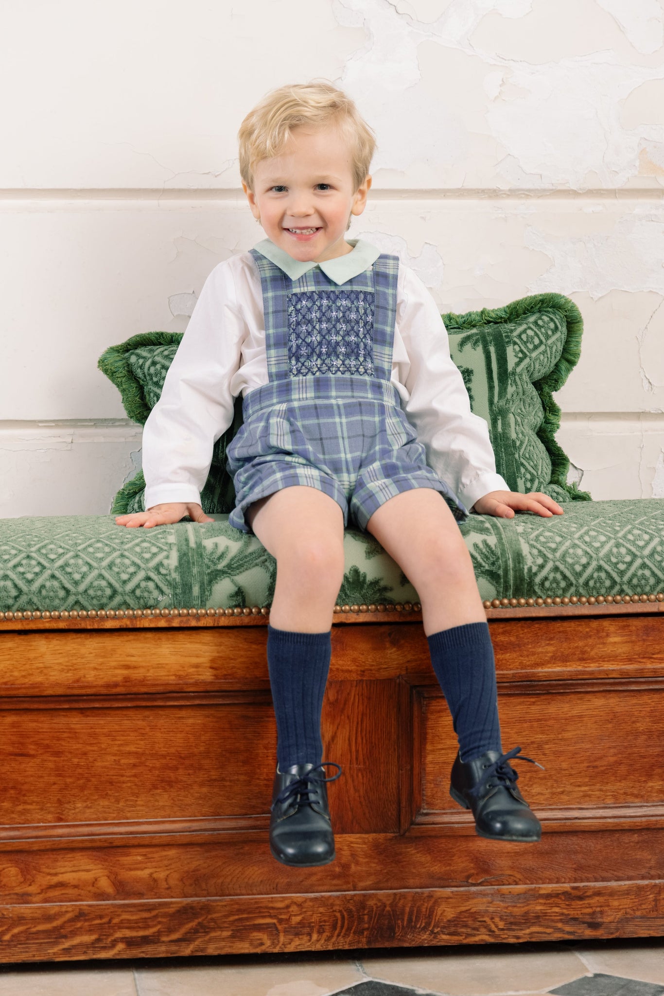 Child wearing George Blue Tartan Overalls and Shirt, sitting on a wooden bench with green cushion.